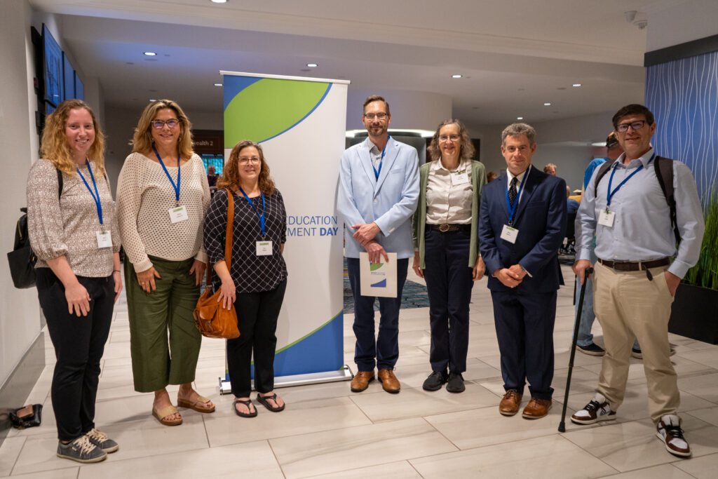 A group of seven people stands indoors in front of an "Education Development Day" banner, all wearing name badges and casual business attire.